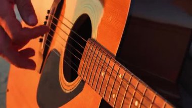 Gün batımında ustalıkla akustik gitar çalan, telleri ve bedeni aydınlatan ellerin yakın çekim videosu. Sıcak ışıklandırma enstrümanın dokusunu ve detaylarını arttırır..