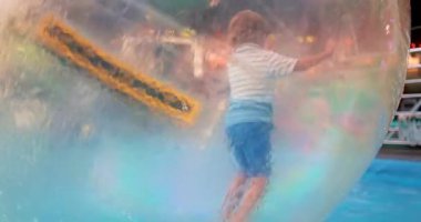 Bir çocuk, lunaparktaki havuzda yüzen şişme bir su topunun içinde oynamaktan zevk alır. Video, bu eşsiz su aktivitesinin heyecanını ve eğlencesini yakalıyor..
