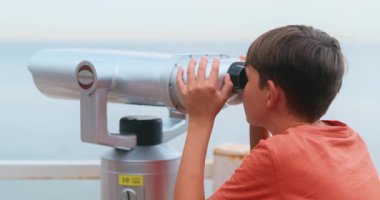 Kırmızı gömlekli bir çocuk, deniz manzaralı bir yere dürbünle bakıyor. Sahne, macera ve keşfin vurgulandığı bir kıyı ortamındaki merak ve keşifleri yakalıyor..