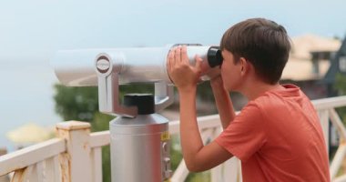 Kırmızı gömlekli genç bir çocuk ahşap bir güverteden uzak manzarayı gözlemlemek için umumi dürbün kullanır. Arkaplanda açık mavi bir gökyüzü ve bulanık yeşillik var. Manzaralı bir bakış açısı var..
