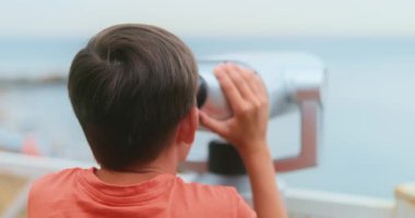 Turuncu gömlekli genç bir çocuk deniz kenarındaki güverteden okyanusu gözlemlemek için dürbün kullanır. Sahne, sakin bir sahil zeminine karşı merak ve keşfi yakalıyor..