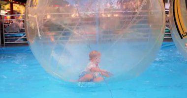 Bir çocuk eğlence parkında suda yüzen şeffaf bir Zorb topunun içinde oynuyor. Sahne, açık hava aktivitelerinin ve aile eğlencesinin coşkusunu yakalıyor..