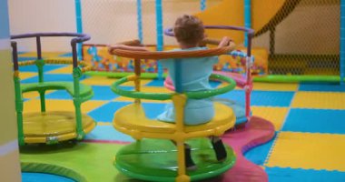 Genç bir çocuk, renkli paspaslar ve güvenlik ağlarıyla çevrili kapalı bir oyun alanındaki canlı oyun parkı ekipmanları üzerinde dönmekten zevk alıyor..