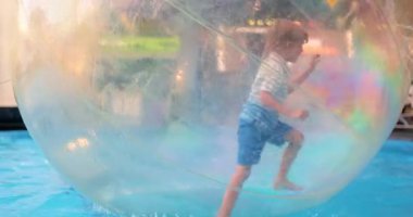 Bir çocuk, lunaparktaki havuzda yüzen şişme bir su topunun içinde oynamaktan zevk alır. Renkli ve şeffaf top eğlenceli ve güvenli su aktivitelerine olanak sağlıyor.