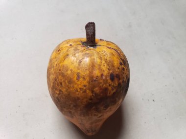 Olgun sapote meyvesinin yakın plan görüntüsü, üzerinde koyu benekler ve küçük bir gövde bulunan sarımsı kahverengi derisini sergiliyor. Meyvenin, pürüzsüz bir yüzeyi ve hafif sivri dibi olan elmaya benzeyen eşsiz bir şekli vardır..