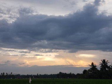 Gün batımında bulutlu bir gökyüzünün manzarası, turuncu ve sarı gölgeler bulutların arasından süzülüyor. Palmiye ağaçlarının siluetleri ön planda görülebiliyor ve yemyeşil pirinç tarlaları araziye yayılıyor..