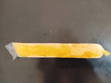 A long, frozen yellow ice pop in a clear plastic wrapper, lying flat on a dark surface. The ice pop has a smooth texture and a tapered end, typical of frozen treats clipart