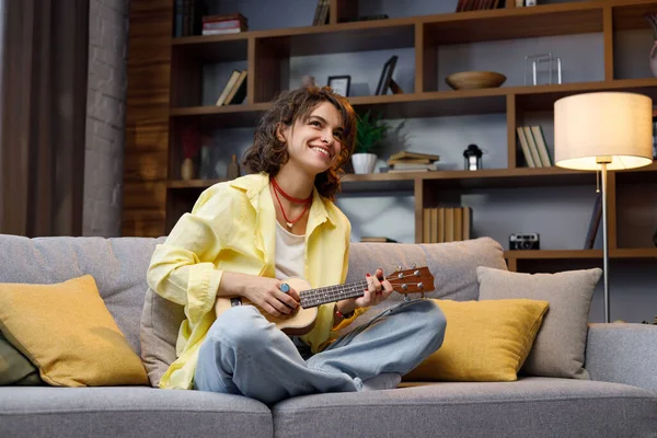 stock image A beautiful hipster girl curly hair in a yellow shirt plays and sings the ukulele on the sofa in the apartment. Home Hobbies playing musical instruments