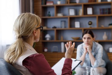 Over shoulder view of female psychologist sitting in armchair, talking with upset woman patient. Psychologist taking notes on clipboard. Psychology, mental therapy, mental health, therapy session.