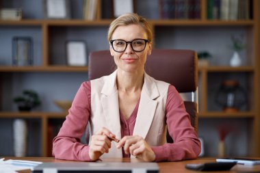 Portrait of beautiful blondie woman looking at camera and smiling. Adult stylish confident businesswoman wearing eyeglasses sitting at workplace in office. Portrait of modern successful female leader.