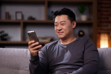 Gülümseyen Asyalı adam sosyal ağlarda iletişim kuruyor ve akıllı telefon kullanıyor. Sıradan giyinmiş bir adam cep telefonundan bakar, dostça iletişim kurar, kanepeye oturur ve evde dinlenir.