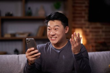 Asyalı adam hareketli başarıyı evet işareti yaparak kutluyor. Heyecanlı Asyalı adam harika haberler alıyor, kazanan jestler yapıyor ve evdeki koltukta otururken telefon ekranına bakıyor.