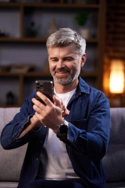 Gülümseyen adam sosyal ağlarda iletişim kuruyor ve akıllı telefon kullanıyor. Sıradan giyinmiş bir adam cep telefonundan bakar, dostça iletişim kurar, kanepeye oturur ve evde dinlenir..