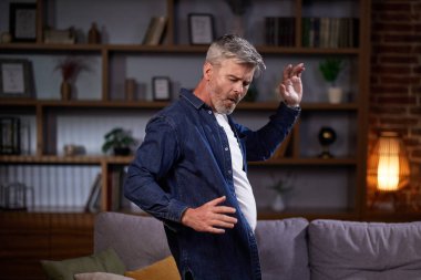 Neşeli adam müzik eşliğinde dans ediyor oturma odasında komik hareketler yapıyor. Günlük kıyafetli bir adam hafta sonu evde kanepenin yanında eğleniyor..