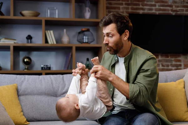 Dad plays with his little baby, on the couch in a cozy apartment. Fatherly love. Father hugs and kisses his little daughter