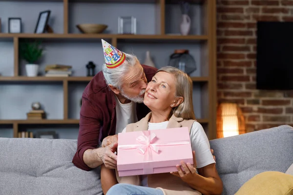Caring old husband giving romantic birthday present to excited mature wife, loving older man surprise happy middle aged woman, open pink box, kiss, hug husband, celebrating together