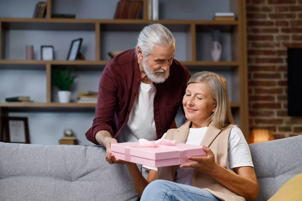Caring old husband giving romantic birthday present to excited mature wife, loving older man surprise happy middle aged woman, open pink box, kiss, hug husband, celebrating together.