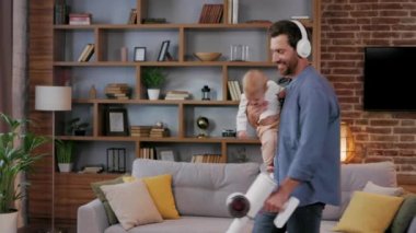 A young father, with a child, does housework. Father on maternity leave. Dad cheerfully sings, dances and vacuums, cleans with a small child in his arms. Paternity Leave.