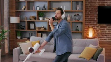 Cheerful guy, the man cleans the apartment. Vacuuming, wearing headphones with dancing and singing into a smartphone instead of a microphone. House cleaning is easy