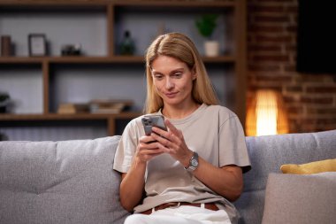 Kanepede oturan sarışın kız akıllı telefona bakıyor, sohbet ediyor, evde dinleniyor. Gülümseyen yetişkin kadın telefonda mesaj yazarak dostça iletişim kurar.