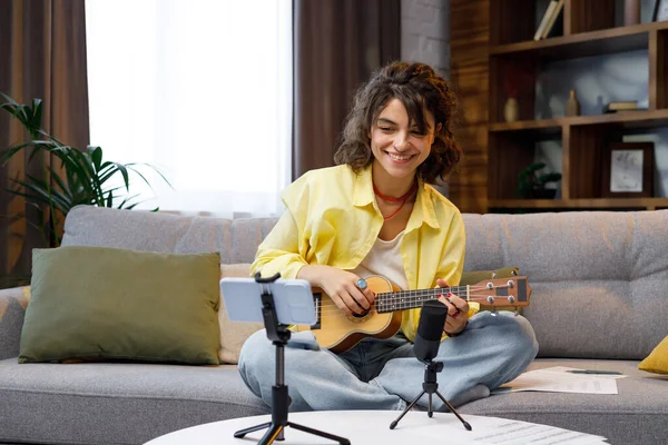 Blogger with mobile phone and microphone. A beautiful hipster girl in a yellow shirt plays and sings on the ukulele on the sofa in the apartment. Online learning to play the guitar