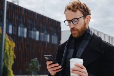 Genç kızıl saçlı bir adam, gözlüklü bir işadamı, şehir tarzının arka planına karşı, akıllı bir telefondan postaları kontrol ediyor, kurye ile iletişim kuruyor ve kahve içiyor. Çevrimiçi iletişim