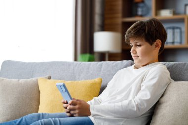 Portrait of young cute boy holding smartphone sitting at sofa. Teenager at home choosing favorite music, texting messages, browsing internet, watching video in social media, playing online games