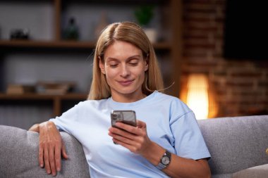 Kanepede oturan sarışın kız akıllı telefona bakıyor, sohbet ediyor, evde dinleniyor. Gülümseyen yetişkin kadın telefonda mesaj yazarak dostça iletişim kurar.