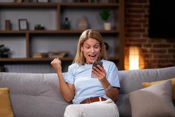 Woman celebrating victory by showing yes gesture on sofa with smartphone. Overjoyed to win, read the good news on the phone and celebrate success at home