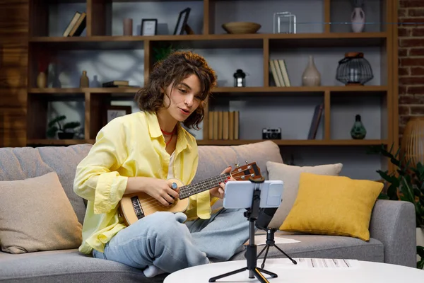 Blogger with mobile phone and microphone. A beautiful hipster girl in a yellow shirt plays and sings on the ukulele on the sofa in the apartment. Online learning to play the guitar