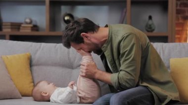 Dad plays with his little baby, on the couch in a cozy apartment. Fatherly love. Father hugs and kisses his little daughter.