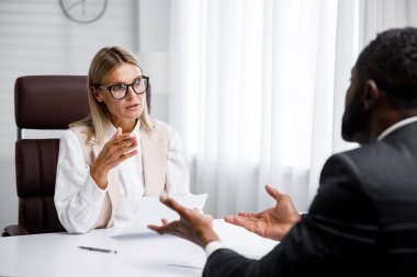 Hiring, interview. Middle-aged blond business woman conducting an interview in a bright office, an African American man. He looks at his resume and after reading it, shakes hands and smiles
