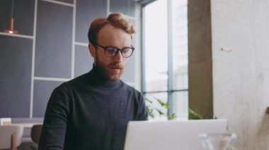 Genç kızıl saçlı bir adam, bir programcı ya da gözlüklü bir girişimci, bir dizüstü bilgisayarın arkasındaki şık bir kafede çalışıyor. Freelancer uzaktan çalışıyor. Çevrimiçi iletişim. Küçük işletme