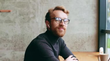 Genç kızıl saçlı bir adam, bir programcı ya da gözlüklü bir girişimci şık bir kafede kameraya bakar ve gülümser. Freelancer uzaktan çalışıyor. Çevrimiçi iletişim. küçük işletme