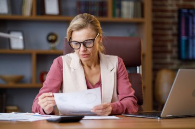 Ofiste çalışan gözlüklü yetişkin bir iş kadınının portresi. Evrak işleriyle uğraşan, mali raporları analiz eden, iş yerinde grafik ve çizelge tutan bir kadın yönetici..