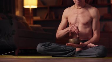 An elderly gray-haired man practices yoga at home using a Tibetan singing bowl for meditation and relaxation