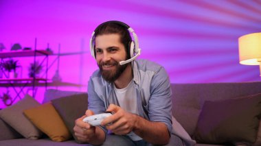 Cheerful young guy in a headset plays video games on the console while sitting on the couch in front of the TV. Gamer having fun together at home. Room with neon light