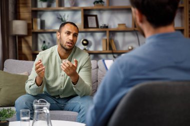 Visiting a psychologist Afro-american man is sitting on the couch and talking to a psychotherapist. He thanks the doctor and smiles, therapy helped him. Help of a psychologist