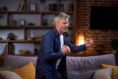 Neşeli adam müzik eşliğinde dans ediyor oturma odasında komik hareketler yapıyor. Günlük kıyafetli bir adam hafta sonu evde kanepenin yanında eğleniyor..