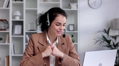 Young curly brunette woman wearing video call headset on laptop in home light office. Video conference, remote meeting. Programmer, remote work, work from home