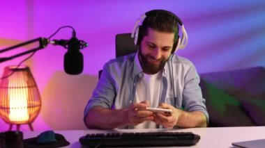 Gamer wearing headphones and headset in a neon room. A bearded guy sits at a table and plays a video game on his smartphone. Mobile video games. Rest and relaxation at home