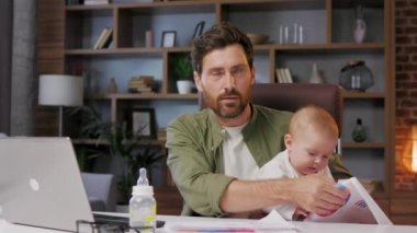 Father is on maternity leave, working in a home office at a laptop with a small child in his arms. Freelance businessman combines child care and work. Paternity Leave