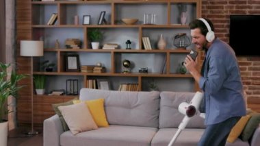Cheerful guy, the man cleans the apartment. Vacuuming, wearing headphones with dancing and singing into a smartphone instead of a microphone. House cleaning is easy
