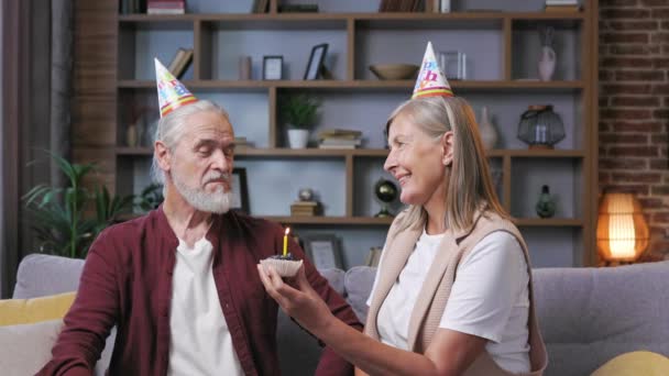 一对头戴圆锥帽的白发苍苍的老夫妇在家里欢度生日 有爱心的丈夫拥抱妻子 庆祝结婚周年 吹灭生日蛋糕上的蜡烛 — 图库视频影像