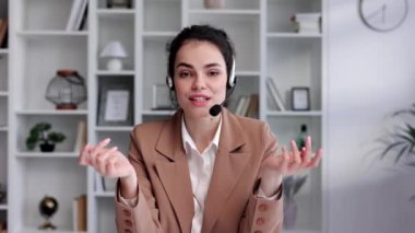 Genç kıvırcık esmer kadın ev beyazının ofisindeki kameradan kulaklık takıyor. Video konferansı, uzaktan görüşme. Programcı, uzaktan çalışma, evden çalışma.