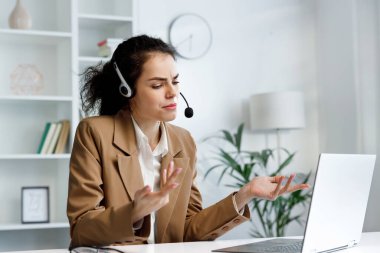 Young curly brunette woman wearing video call headset on laptop in home light office. Video conference, remote meeting. Programmer, remote work, work from home