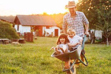 Küçük mutlu çocuklar, sıcak ve güneşli bir günde evcil bahçede babalarıyla el arabasında eğleniyorlar. Çocuklu aileler için açık hava oyunları. Hafta sonları kırsalda birlikte vakit geçirmek.