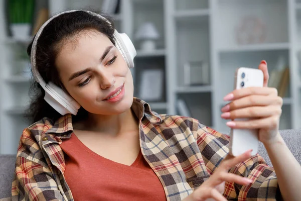 Young student girl in white headphones sitting on the sofa and taking a selfie in a beautiful bright apartment