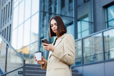 A young brunette girl against the backdrop of a business center, office center. Portrait of a successful startup. Smartphone, coffee casual suit