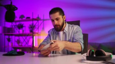 Gamer in neon room. A bearded guy sits at a table and plays a video game on his smartphone. Mobile video games. Rest and relaxation at home